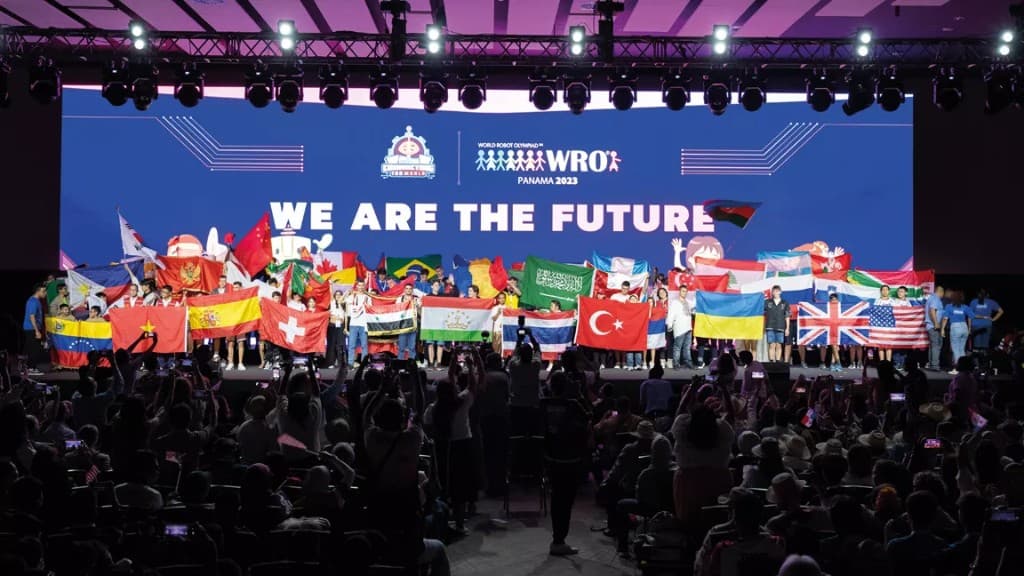 International World Robot Olympiad stage with teams, national flags, and a WE ARE THE FUTURE headline on screen.